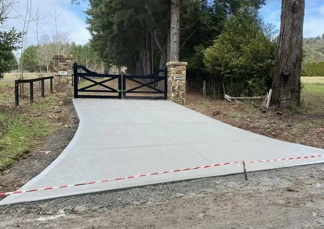 A plain concrete driveway leading up to gates in a rural setting. | DigKing